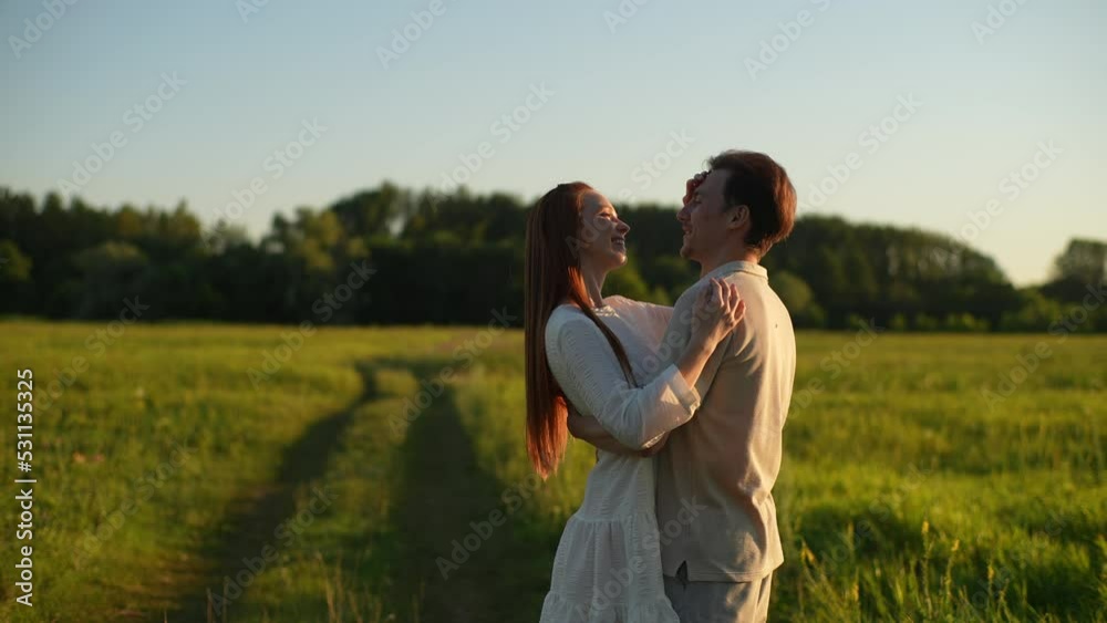Portrait of loving young man and redhead woman in love hugging, kissing, stroking standing on field with green grass, enjoying each other, on background of sunlight in summer evening during sunset. Portrait of loving young man and redhead woman in love hugging, kissing, stroking standing on field with green grass, enjoying each other, on background of sunlight in summer evening during sunset.