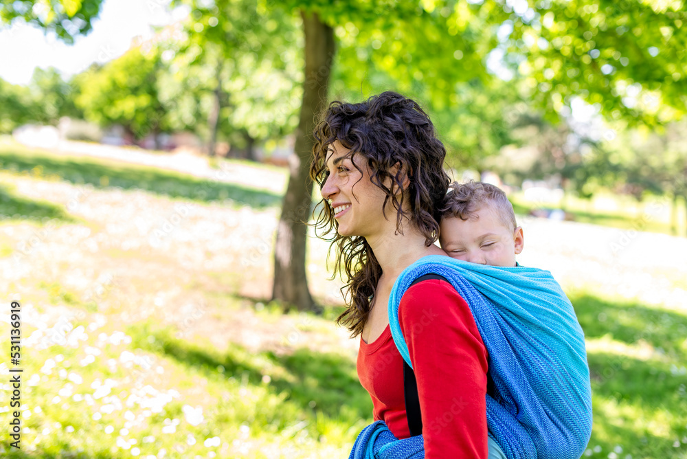 Fototapeta premium close up portrait of a happy young mom carries a child on her back tied up with a colorful traditional piece of fabric in a public park garden. mother and son in a baby wrap sling chilling outdoor.