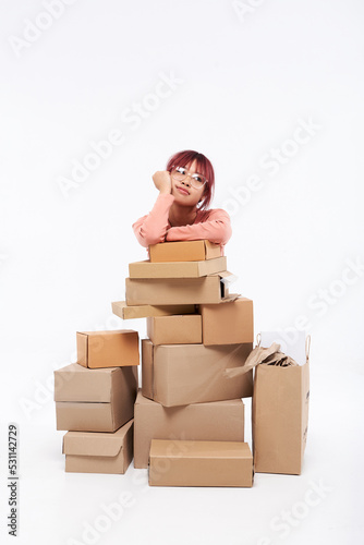 Portrait of a beautiful young Asian red hair brunette in glasses sitting under many boxes and think to move into her new home. Moving stuff. Resettlement, packaging or delivery concept. Copy space
