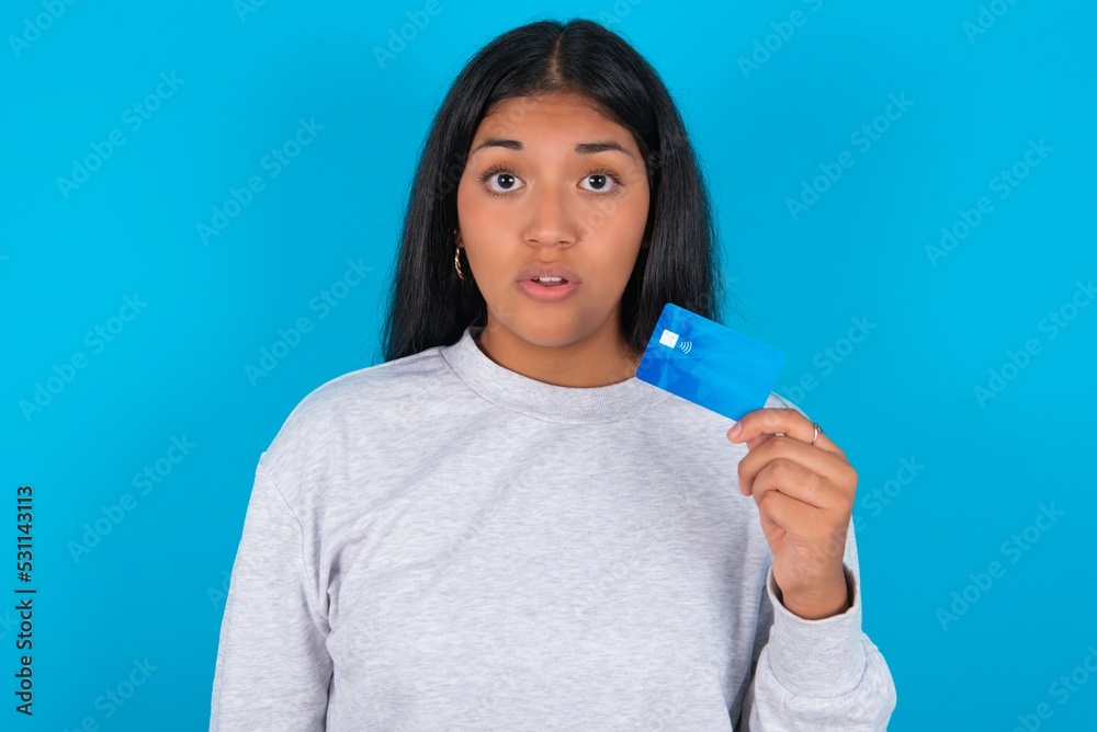 Photo of Young hispanic woman wearing grey sweatshirt over blue background amazed shocked hold credit card payment Photo of Young hispanic woman wearing grey sweatshirt over blue background amazed shocked hold credit card payment