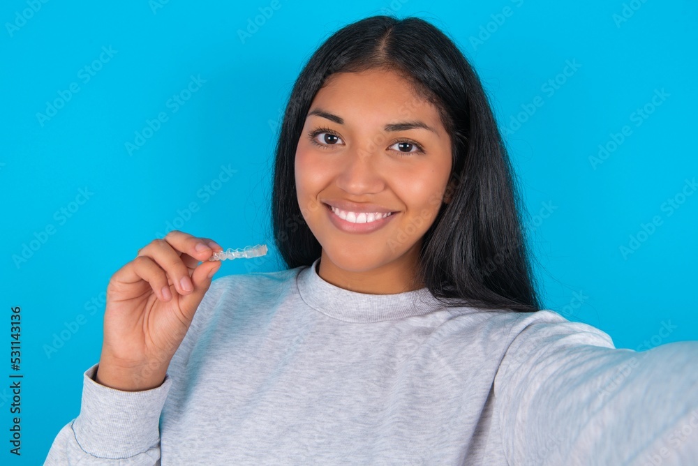 Young hispanic woman wearing grey sweatshirt over blue background make selfie holding an invisible braces aligner, recommending this new treatment. Dental healthcare concept. Young hispanic woman wearing grey sweatshirt over blue background make selfie holding an invisible braces aligner, recommending this new treatment. Dental healthcare concept.