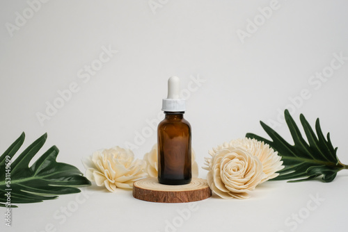 amber glass bottle with dropper on wooden podium with flower and green leaf. front view.