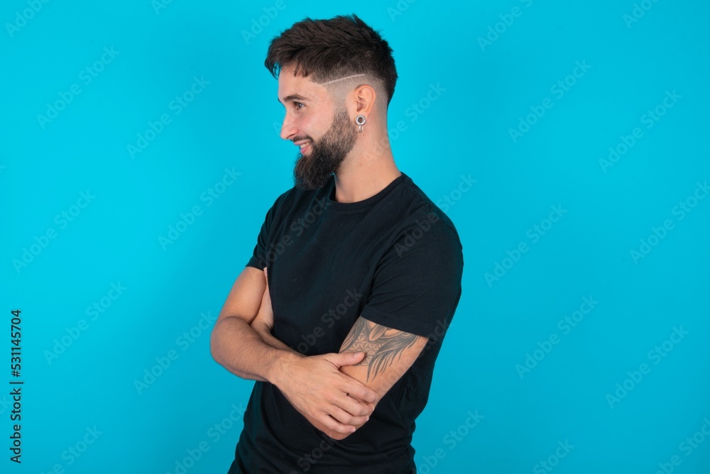 young hispanic bearded man wearing black T-shirt standing against blue background cross hands look copy space young hispanic bearded man wearing black T-shirt standing against blue background cross hands look copy space