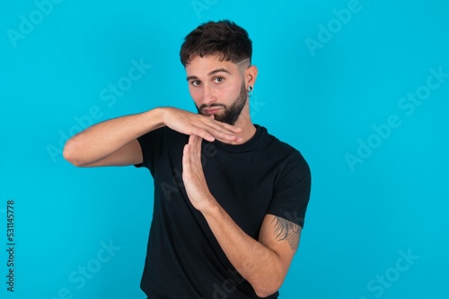 young hispanic bearded man wearing black T-shirt standing against blue background being upset showing a timeout gesture, needs stop, asks time for rest after hard work,