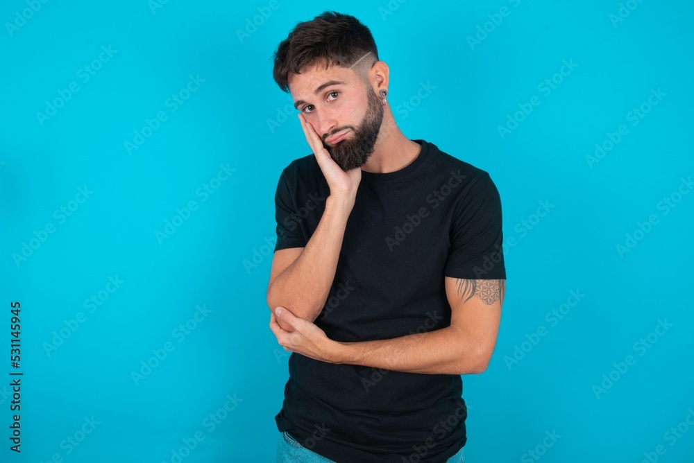 Very bored young hispanic bearded man wearing black T-shirt standing against blue background holding hand on cheek while support it with another crossed hand, looking tired and sick, Very bored young hispanic bearded man wearing black T-shirt standing against blue background holding hand on cheek while support it with another crossed hand, looking tired and sick,