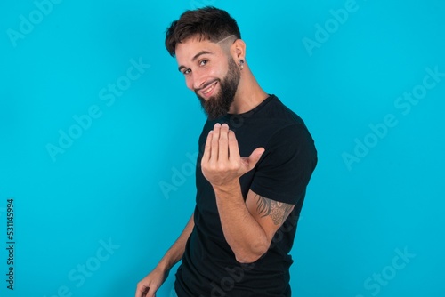 young hispanic bearded man wearing black T-shirt standing against blue background inviting to come with hand. Happy that you came photo