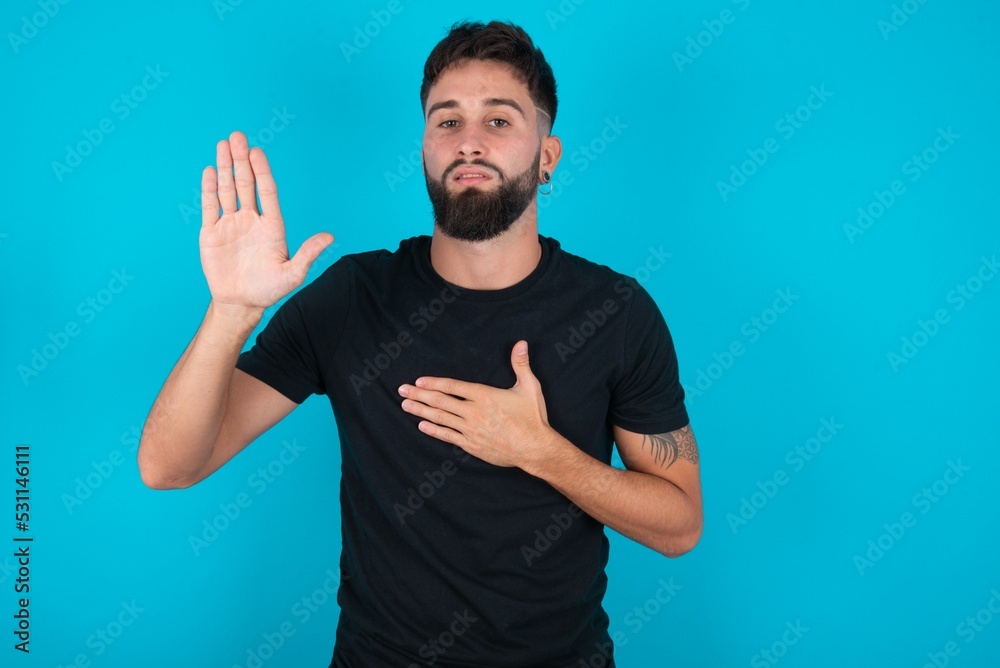 young hispanic bearded man wearing black T-shirt standing against blue background Swearing with hand on chest and open palm, making a loyalty promise oath young hispanic bearded man wearing black T-shirt standing against blue background Swearing with hand on chest and open palm, making a loyalty promise oath