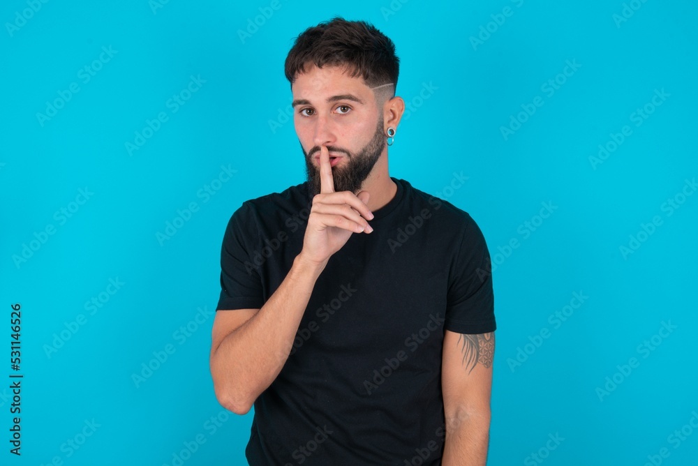 young bearded hispanic man wearing black T-shirt over blue background makes silence gesture, keeps finger over lips. Silence and secret concept. young bearded hispanic man wearing black T-shirt over blue background makes silence gesture, keeps finger over lips. Silence and secret concept.