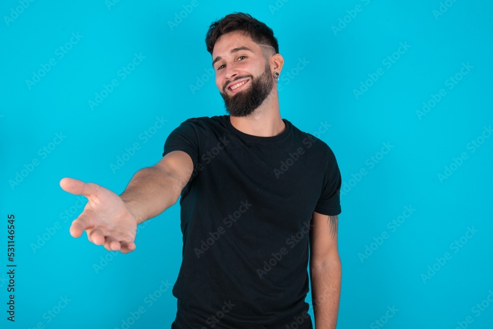 young bearded hispanic man wearing black T-shirt over blue background smiling friendly offering handshake as greeting and welcoming. Successful business.