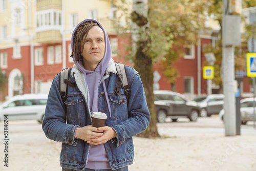 Young handsome man with dreadlocks in warm clothes holding paper cup with coffee, standing in city street. © Anton Dios