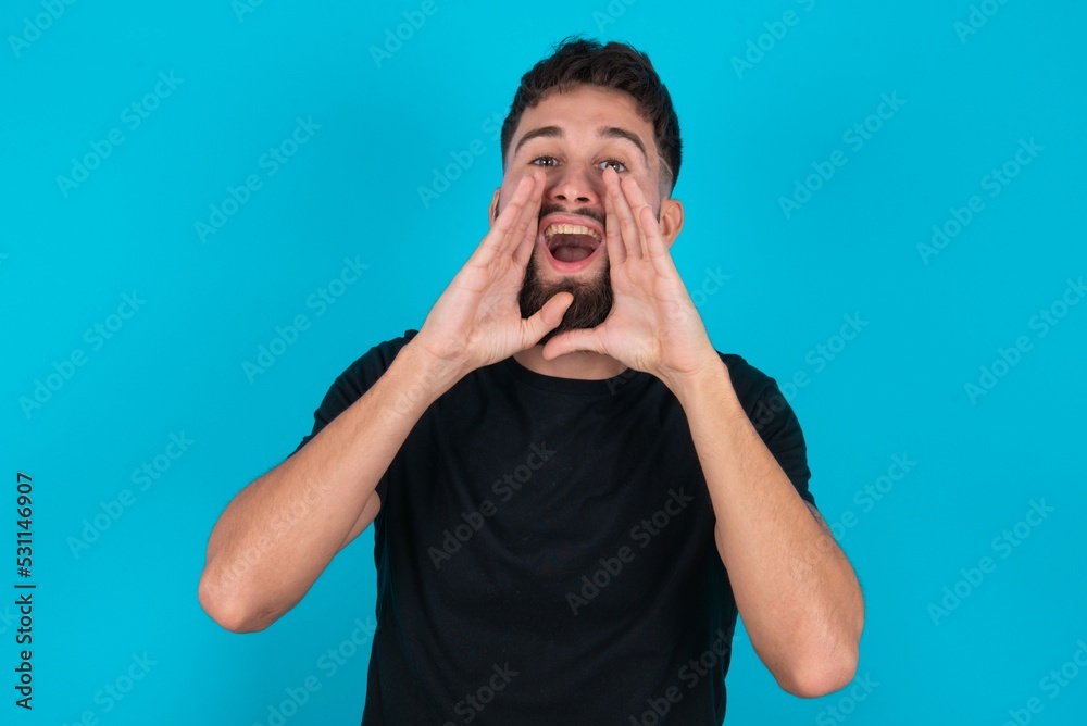 young bearded hispanic man wearing black T-shirt over blue background shouting excited to front. young bearded hispanic man wearing black T-shirt over blue background shouting excited to front.