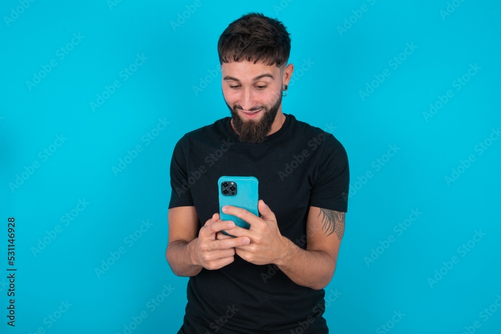 young bearded hispanic man wearing black T-shirt over blue background using mobile phone chatting free time . young bearded hispanic man wearing black T-shirt over blue background using mobile phone chatting free time .
