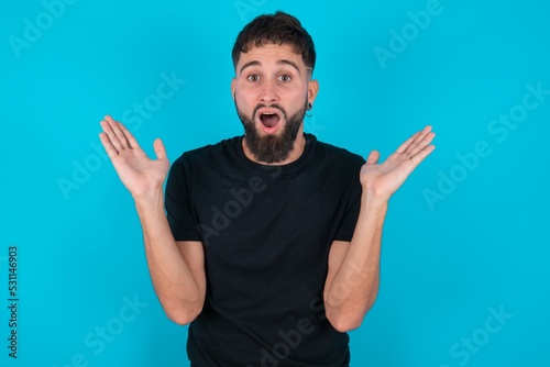 Surprised terrified young bearded hispanic man wearing black Gestures with uncertainty, stares at camera, puzzled as doesn't know answer on tricky question, People, body language, emotions concept