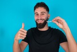 young bearded hispanic man wearing black T-shirt over blue background holding an invisible braces aligner and rising thumb up, recommending this new treatment. Dental healthcare concept.