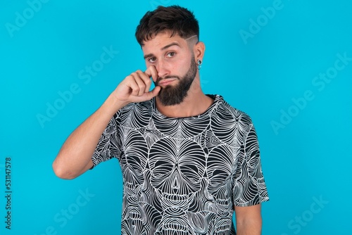 Disappointed dejected young bearded hispanic man wearing casual clothes over blue background wipes tears stands stressed with gloomy expression. Negative emotion