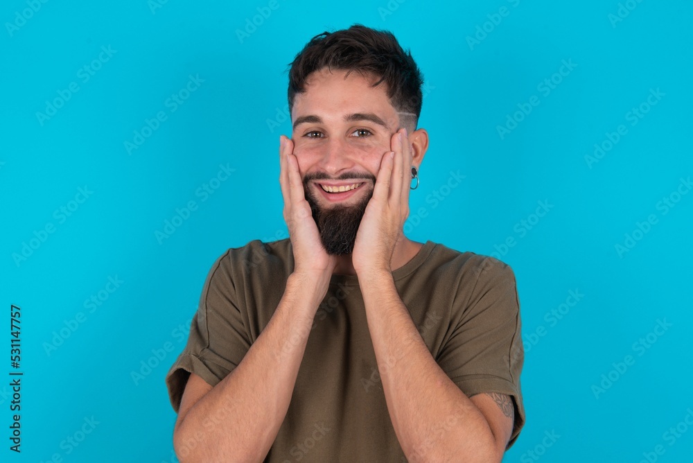 Happy young bearded hispanic man wearing casual clothes over blue background touches both cheeks gently, has tender smile, shows white teeth, gazes positively straightly at camera, Happy young bearded hispanic man wearing casual clothes over blue background touches both cheeks gently, has tender smile, shows white teeth, gazes positively straightly at camera,