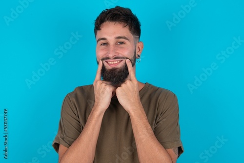 Happy young bearded hispanic man wearing casual clothes over blue background with toothy smile, keeps index fingers near mouth, fingers pointing and forcing cheerful smile