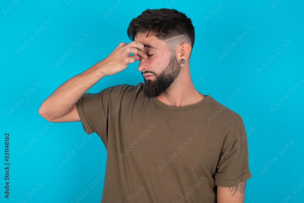 Sad young bearded hispanic man wearing casual clothes over blue background suffering from headache holding hand on her face Sad young bearded hispanic man wearing casual clothes over blue background suffering from headache holding hand on her face