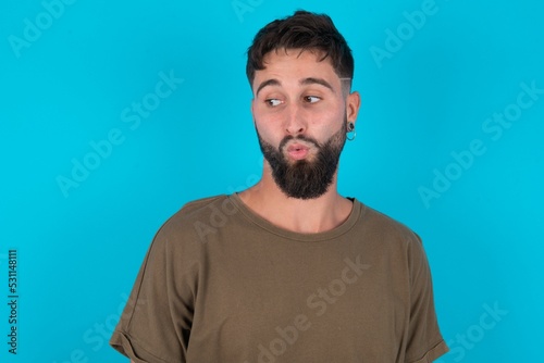 Shocked young bearded hispanic man wearing casual clothes over blue background look empty space with open mouth screaming: Oh My God! I can't believe this.