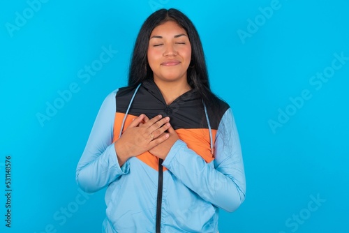 young beautiful woman wearing sport outfit  closes eyes and keeps hands on chest near heart, expresses sincere emotions, being kind hearted and honest. Body language and real feelings concept. photo