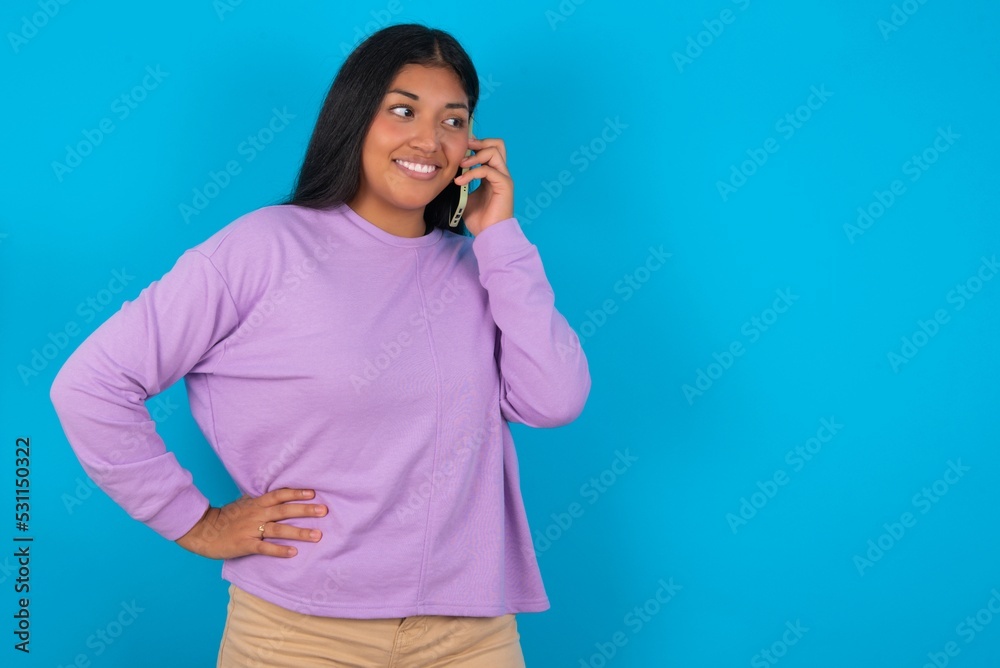 Portrait of successful joyful young beautiful latin woman wearing casual clothes standing against blue background talking on mobile phone with friend. Lifestyle and communication concept Portrait of successful joyful young beautiful latin woman wearing casual clothes standing against blue background talking on mobile phone with friend. Lifestyle and communication concept