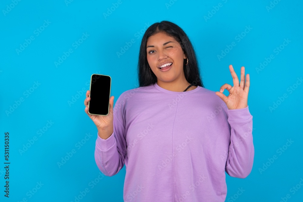 Excited young beautiful latin woman wearing casual clothes standing against blue background showing smartphone blank screen, blinking eye and doing ok sign with hand. Advertisement concept. Excited young beautiful latin woman wearing casual clothes standing against blue background showing smartphone blank screen, blinking eye and doing ok sign with hand. Advertisement concept.
