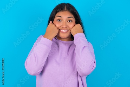 Happy Young beautiful hispanic woman wearing casual clothes over blue background keeps fists on cheeks smiles broadly and has positive expression being in good mood