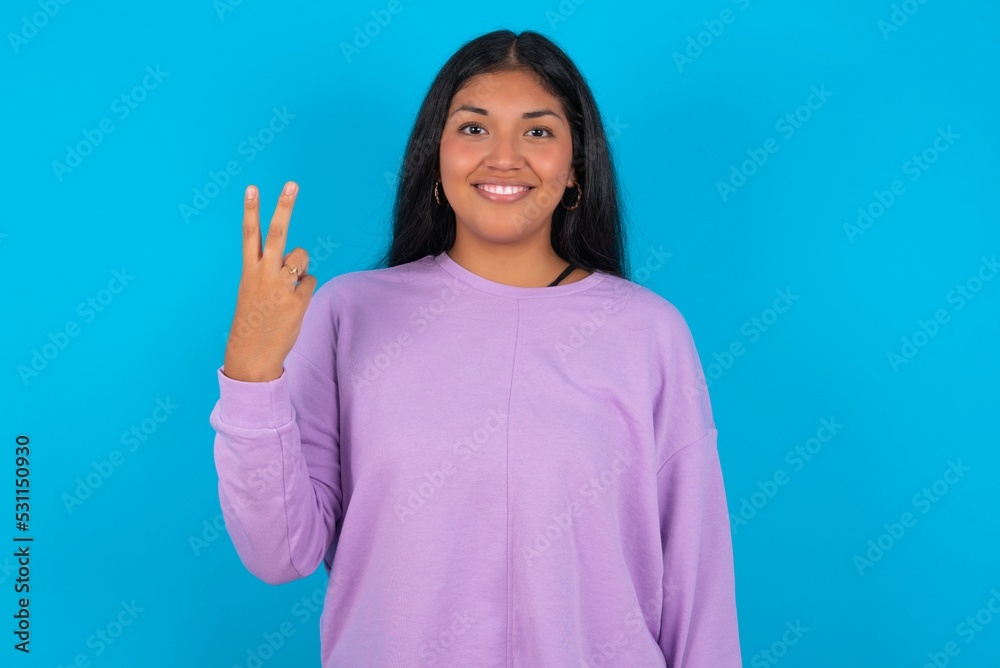 Young beautiful hispanic woman wearing casual clothes over blue background smiling and looking friendly, showing number two or second with hand forward, counting down Young beautiful hispanic woman wearing casual clothes over blue background smiling and looking friendly, showing number two or second with hand forward, counting down