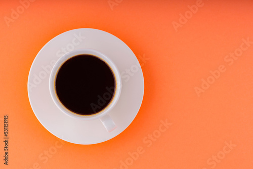 Cup of coffee on orange background. White cup of black coffee on a white saucer. Copy space. Top view