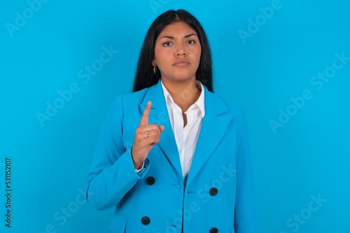 Young latin businesswoman wearing blue blazer over blue background frustrated and pointing to the front
