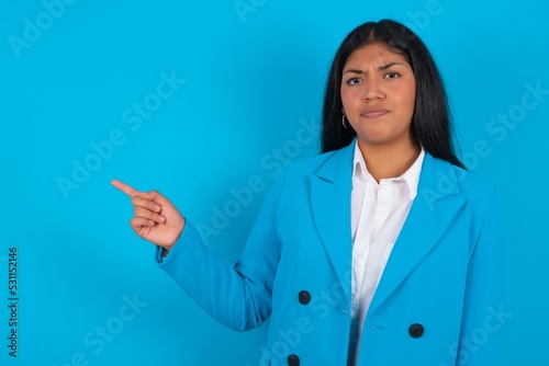 Serious Young latin businesswoman wearing blue blazer over blue background smirks face points away on copy space shows something unpleasant. Look at this advertisement. Big price concept