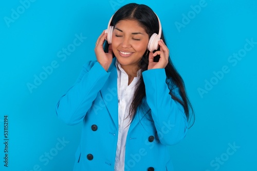 Pleased Young latin businesswoman wearing blue blazer over blue background enjoys listening pleasant melody keeps hands on stereo headphones closes eyes. Spending free time with music