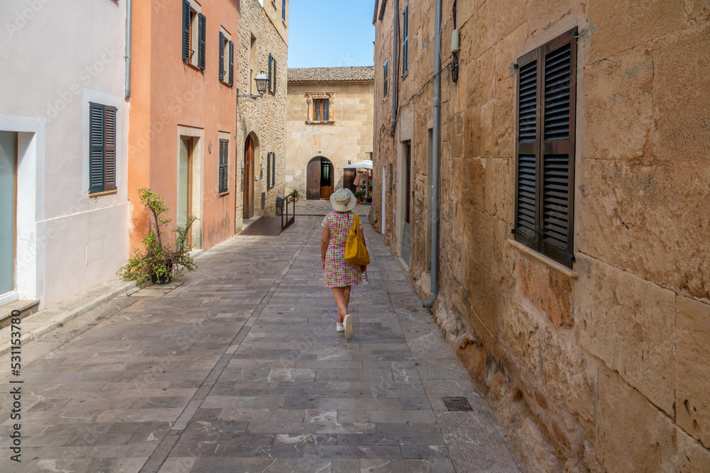Woman seen from behind, with a hat, bag and colorful dress, walking along a typical street in the Majorcan town of Alcudia on a sunny summer morning