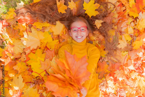 a teenage girl in red glasses lies in yellow autumn foliage and smiles