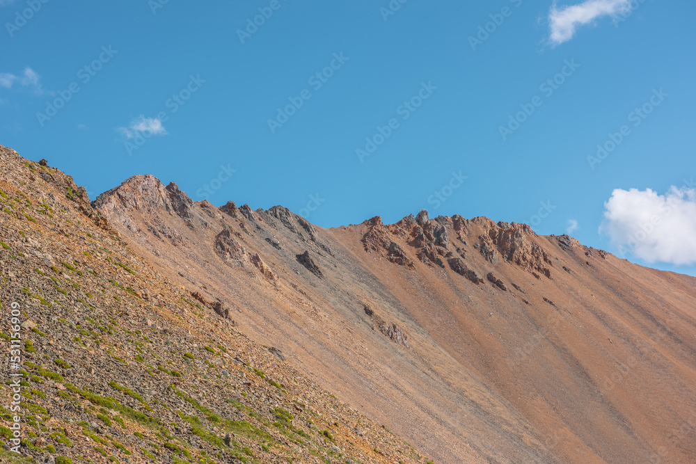 Scenic landscape with sunlit high rocky mountain ridge under blue sky in sunny day. Colorful scenery with large mountain wall in sunlight. Scenic view to sharp rocks on mountain top in bright sun.