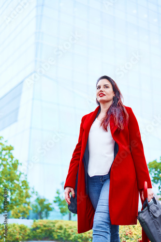 Middle-aged woman walking to a job interview dressed in a white blouse and red coat looking to the side. Defocused background of an office building and the sky. Copy space Business executive woman.