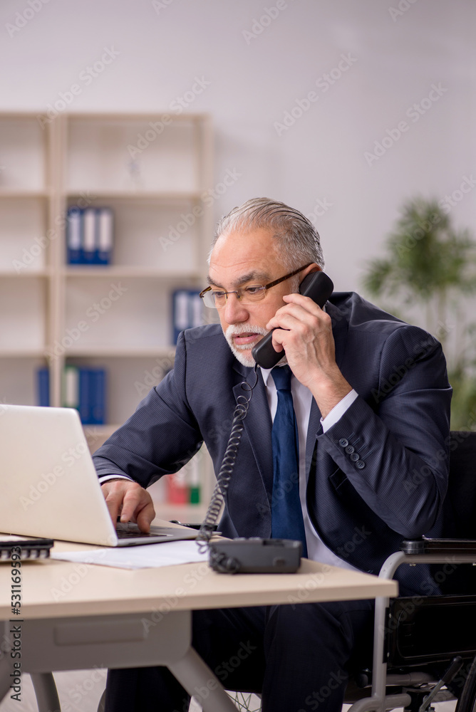 Old male employee in wheel-chair sitting at workplace Old male employee in wheel-chair sitting at workplace