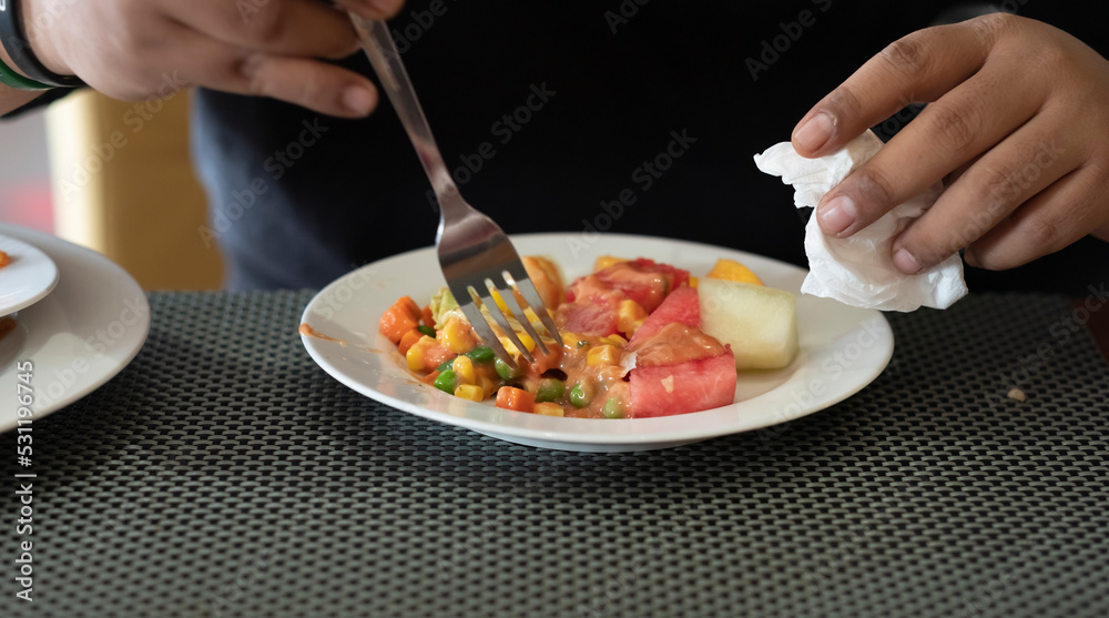 Cropped image of a young casual man eating fresh vegetables and fruits for breakfast. Right hand is holding a fork and left hand is holding a tissue.