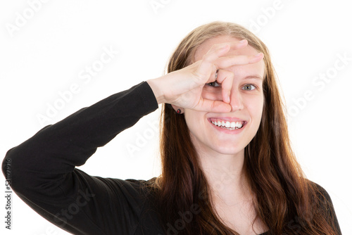 Joyful beautiful woman with open mouth applying fingers hand to eye and making Ok gesture on white background