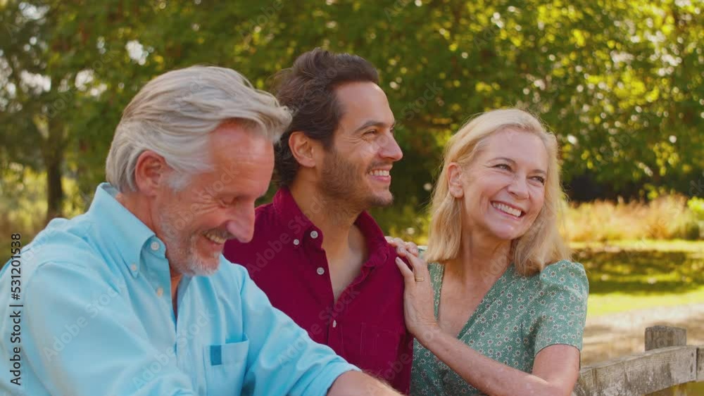 Smiling mature parents with mixed race adult son leaning on wooden fence on walk in summer countryside talking together - shot in slow motion Smiling mature parents with mixed race adult son leaning on wooden fence on walk in summer countryside talking together - shot in slow motion