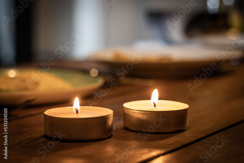 two lit candles on top of a brown wooden table, with plate in the background