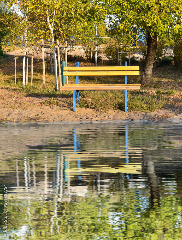 Fototapeta premium One yellow bench in the autumn park is reflected in the water. Beautiful autumn background. Loneliness concept.