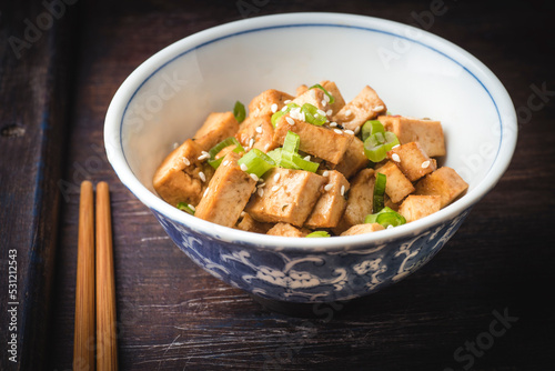 Autumn hot Japanese recipe. Fried tofu with sesame seeds, green onion and spices on ceramic plate with chopsticks, wooden vintage background. Homemade healthy vegetarian Asia and Japanese dish