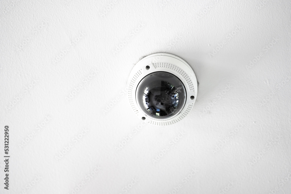 White CCTV with acrylic glass dome cover camera len in the centre hanging from the ceiling. The background is white studio. White CCTV with acrylic glass dome cover camera len in the centre hanging from the ceiling. The background is white studio.