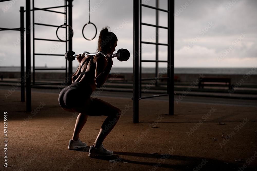 young athletic woman standing on bent knees and holding barbell on her shoulders. - obrazy, fototapety, plakaty 