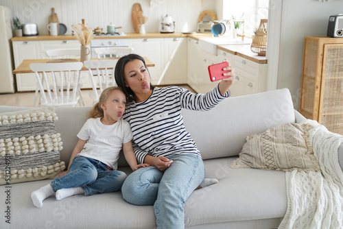 Little daughter, mom show tongues, take selfie photo by smartphone, sitting on sofa together at home