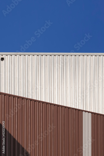 Sunlight on surface of 2 brown and silver corrugated steel warehouse building against blue clear sky in vertical frame