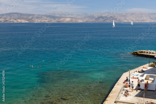 Bathing in Corfu town, blue water. In the background, a mountain range.