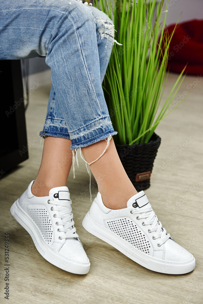 Young woman in blue jeans trying on white sport shoes in shop, copy space. Woman wearing comfortable stylish sneakers indoors, closeup Young woman in blue jeans trying on white sport shoes in shop, copy space. Woman wearing comfortable stylish sneakers indoors, closeup
