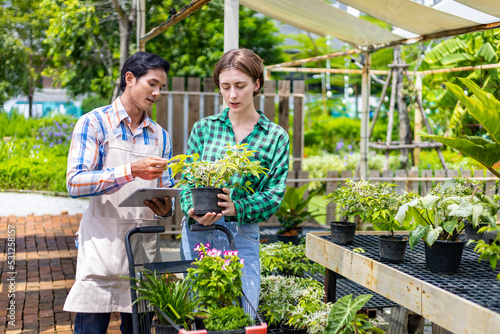 Young caucasian customer is asking the nursery owner about the exotic plant with shopping cart full of summer plant for weekend gardening and outdoor pursuit